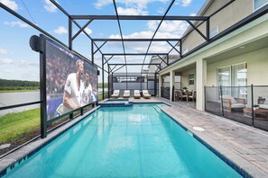 A swimming pool with lounge chairs, a covered seating area, and a large screen displaying a sports event. - 7BR house sleeps 20 with hot tub and pool (Kissimmee)
