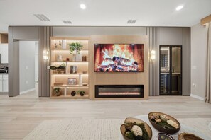 A modern living room with a large flat-screen TV mounted on a wooden wall, a built-in fireplace, and a decorative shelf with plants and books. - 7BR house at Windsor Cay Resort near Disney (Clermont)