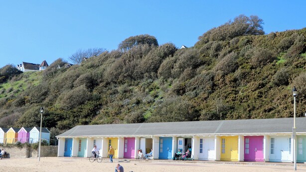 A row of colorful beach huts with a sandy beach in the foreground and a hillside in the background. - Woodlands House (Ringwood)