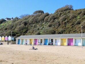 A row of colorful beach huts with a sandy beach in the foreground and a hillside in the background.