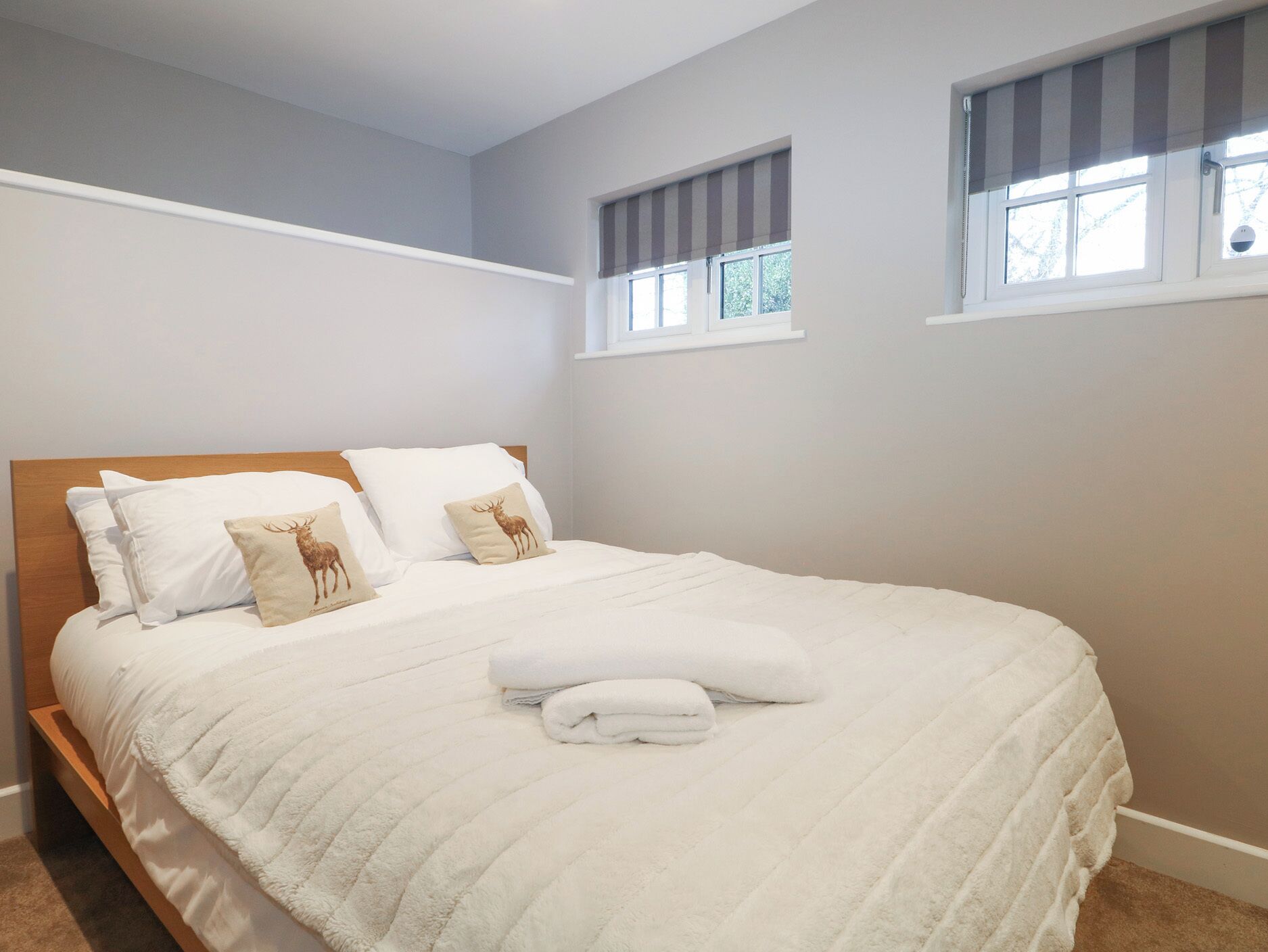 Een netjes opgemaakt bed met witte lakens en twee kussens, waarvan één met een hertenprint, in een kamer met lichtgrijze wanden en een raam met een gestreept rolgordijn.