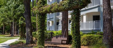 Un bâtiment doté d’un espace extérieur aménagé avec des sièges, protégé par un toit, et entouré d’arbres et d’un sentier.