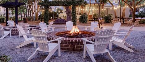 A fire pit surrounded by white Adirondack chairs in an outdoor setting with trees and a building in the background.