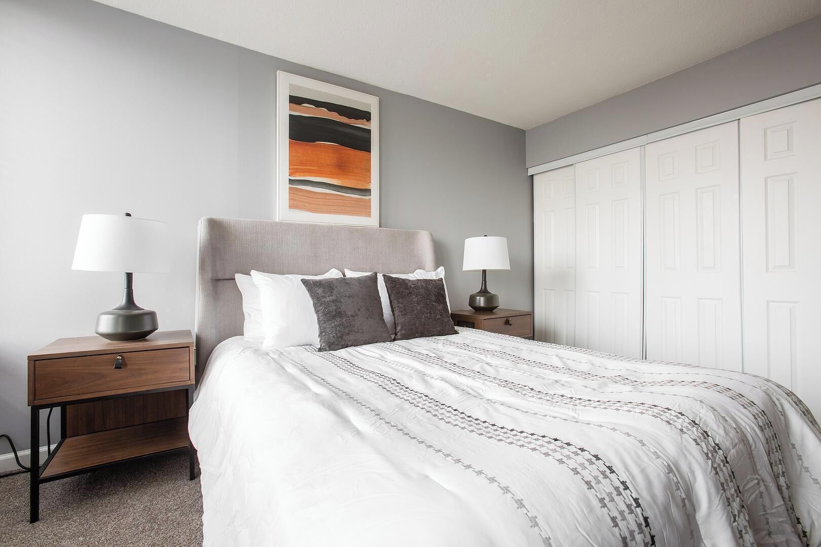 A bedroom with a bed, two bedside tables with lamps, a wardrobe, and a framed abstract painting on the wall.