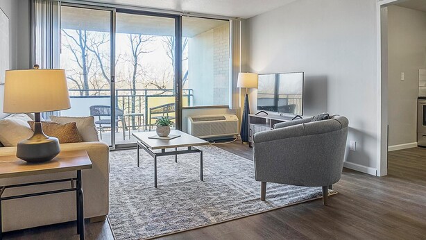 A modern living room with a sofa, a coffee table, a TV, and a large window with a view of trees. - Landing Apartments Fort Wayne - East Central (Fort Wayne)