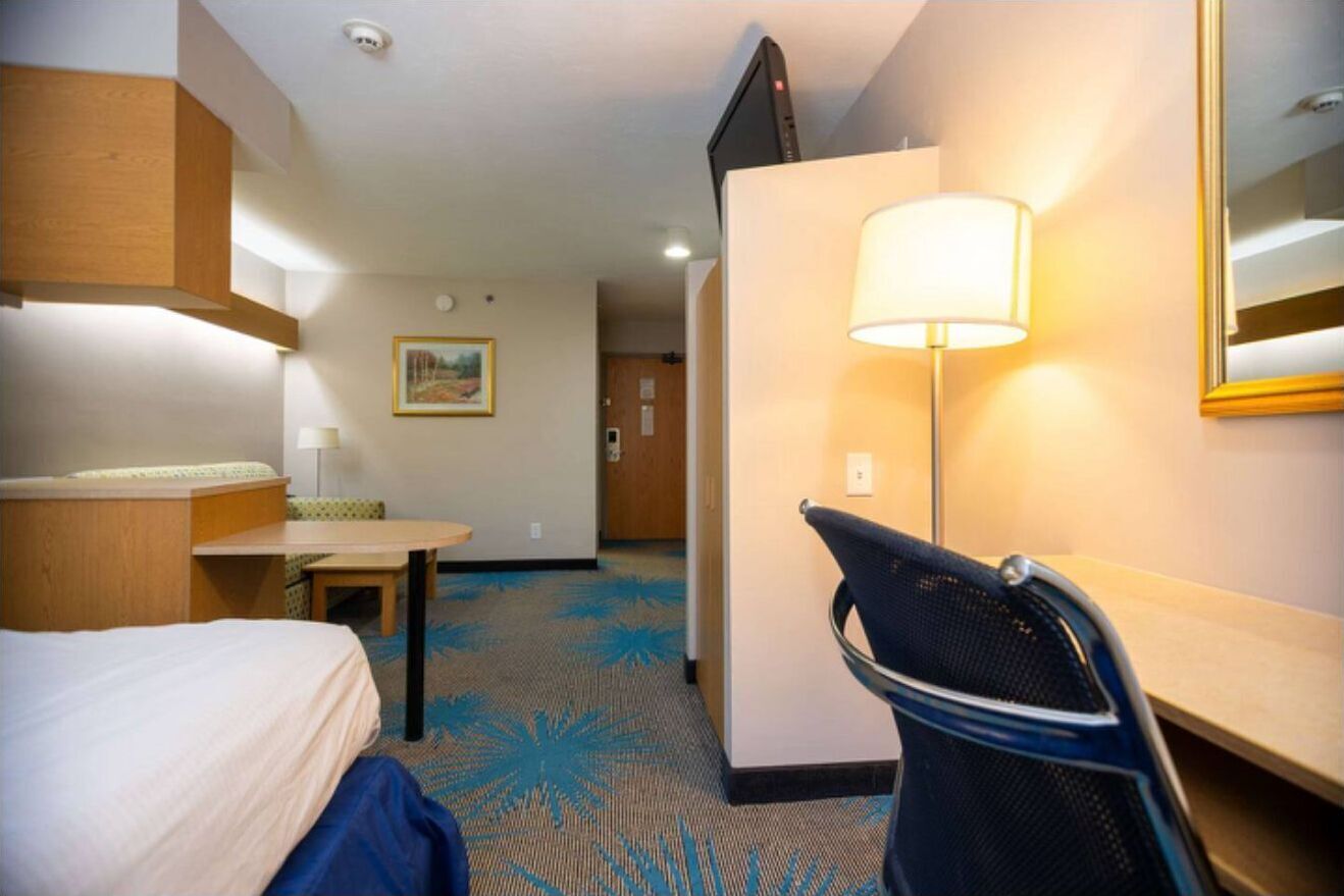 A hotel room with a bed, a desk, a chair, a lamp, and a mirror.