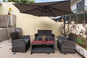 Een terras buiten met rieten meubels, een tafel met een fles en glazen, en een grote parasol.