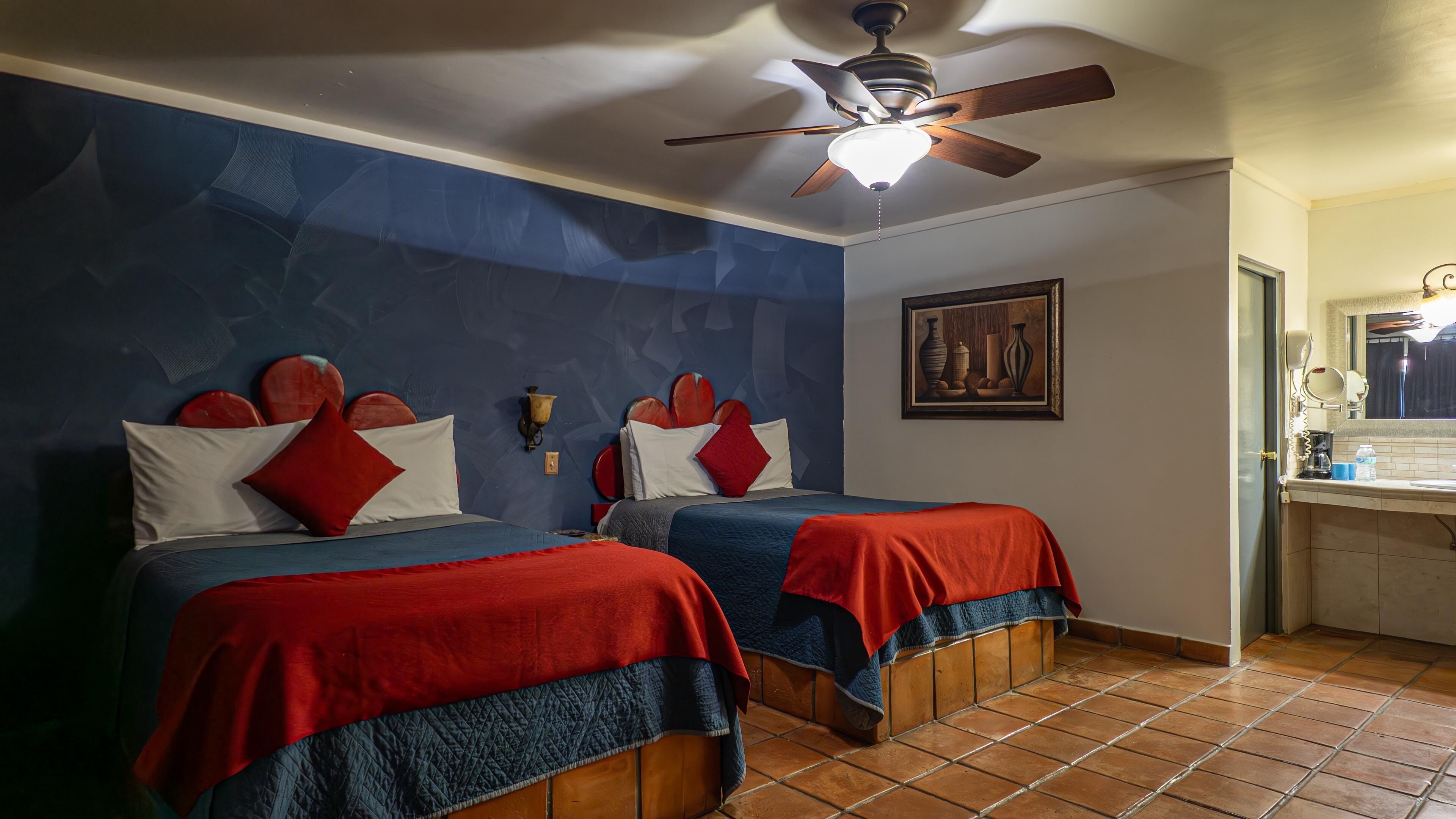 Habitación de hotel con dos camas, ventilador de techo, un cuadro y baño con espejo y lavamanos.