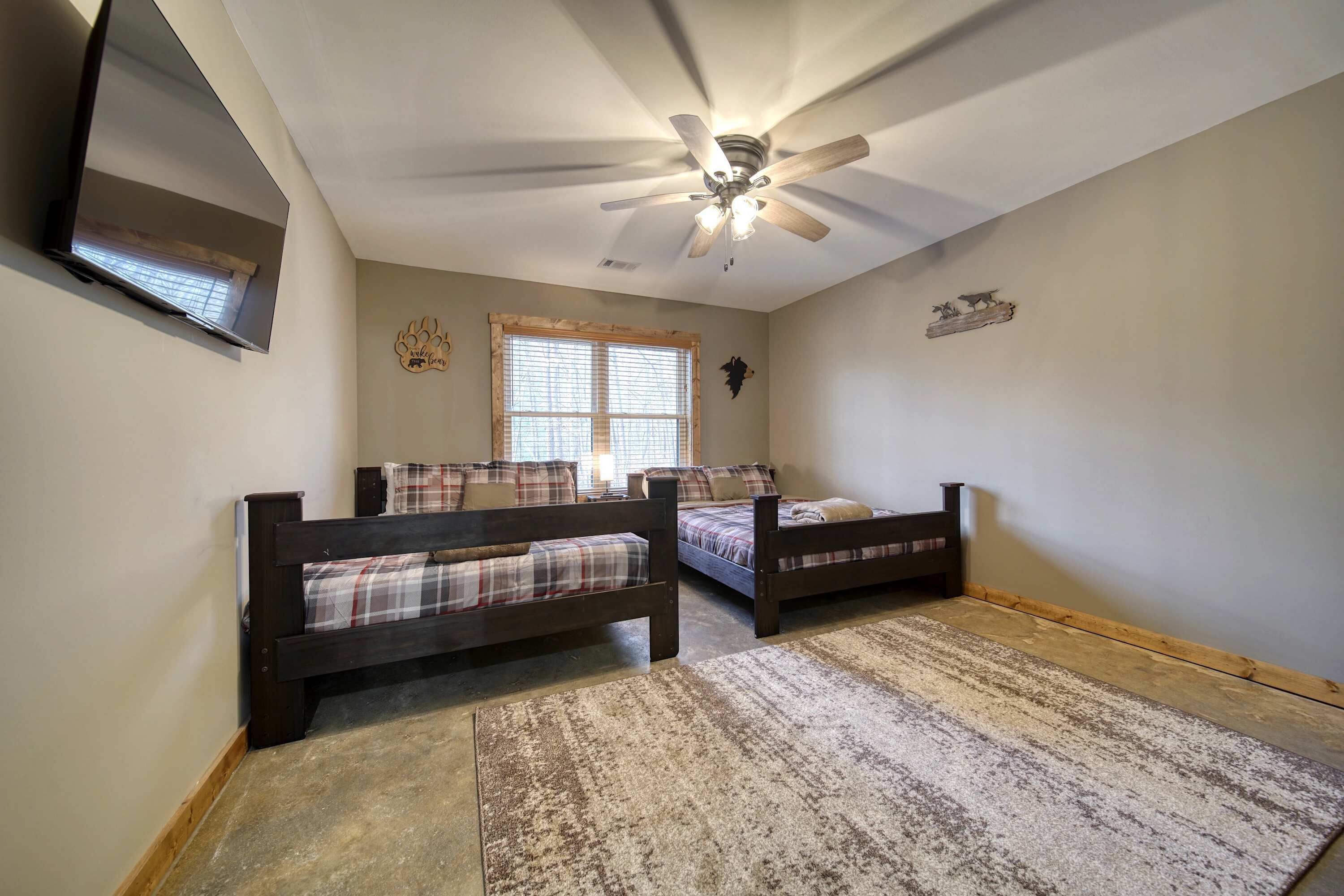 A bedroom with two wooden beds, a ceiling fan, a TV mounted on the wall, and a window with blinds.
