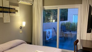 A hotel room with a large bed, a desk, a chair, an air conditioning unit, a wall-mounted TV, and a sliding glass door leading to an outdoor area with a table and chairs.