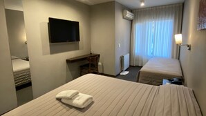 A hotel room with two beds, a desk, a flat-screen TV, an air conditioning unit, and a window with curtains.