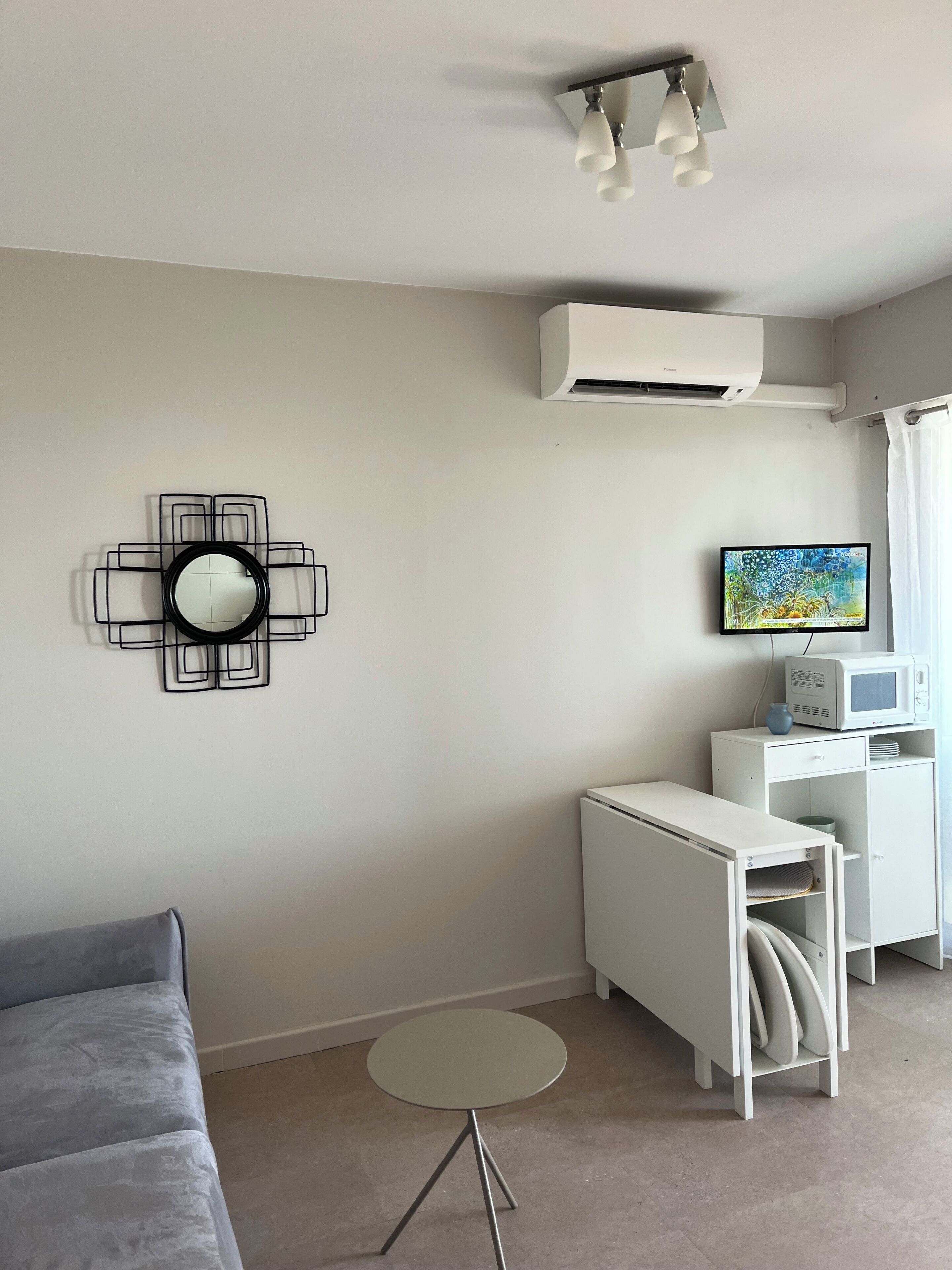 Un salon moderne avec un canapé gris, une petite table ronde, un meuble blanc, un téléviseur à écran plat et un climatiseur fixé au mur.