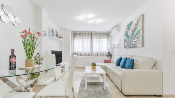 Una sala de estar moderna con una mesa de comedor de vidrio, sofás blancos y un amplio ventanal con cortinas de gasa.
