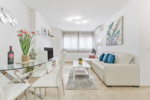 Una sala de estar moderna con una mesa de comedor de vidrio, sofás blancos y un amplio ventanal con cortinas de gasa.