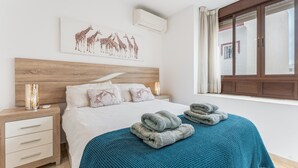 Un dormitorio con cama, una mesita de noche, un cuadro de jirafas en la pared, una ventana con cortinas y una manta azul sobre la cama.