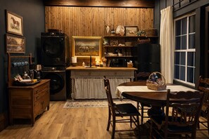 A cozy kitchen with wooden cabinets, a dining table, and chairs. There are framed pictures on the wall, a coffee machine, and a microwave.