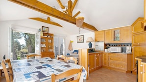 Une cuisine avec des meubles en bois, une table à manger recouverte d’une nappe à motifs, et une vue sur l’extérieur par la porte ouverte.