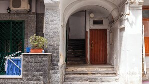Uma escadaria estreita leva a uma porta de madeira, limitada por paredes de pedra e um vaso de planta.