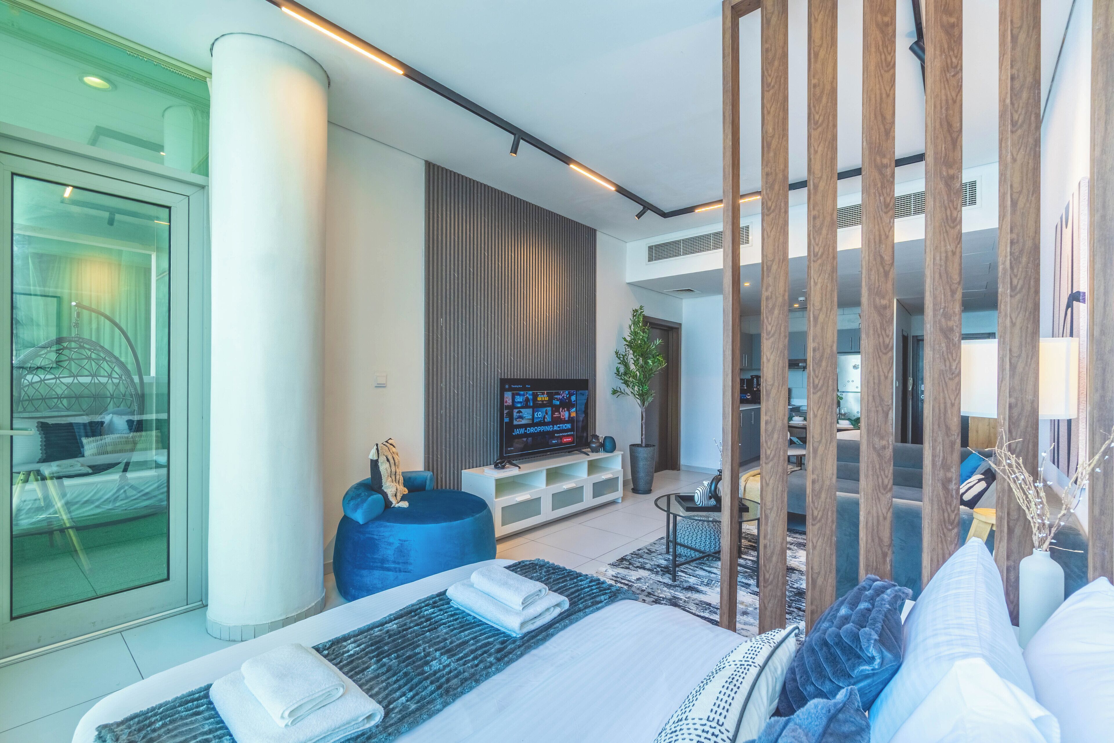 Une chambre d’hôtel moderne, dotée d’un grand lit, d’une télévision, d’un canapé bleu et d’un coin repas visible à travers une porte vitrée.