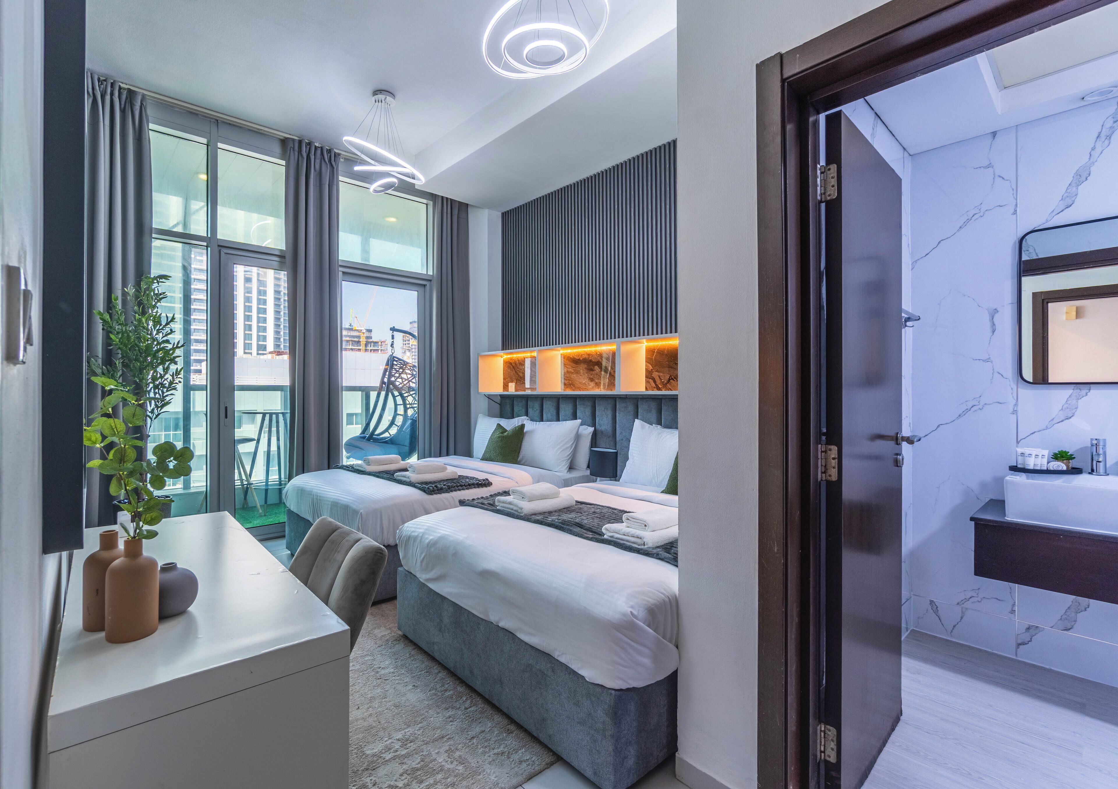 A modern hotel room with two beds, a desk, and a large window with a view of the city.