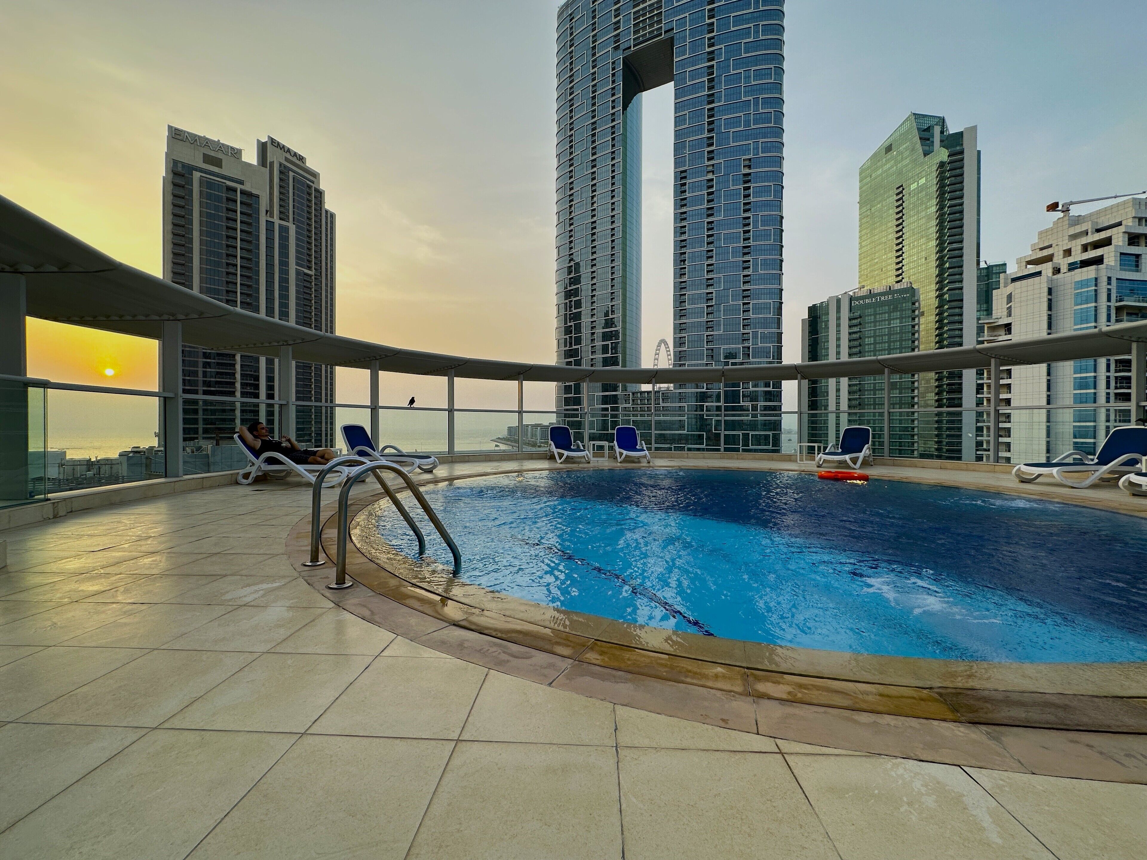 A rooftop pool with lounge chairs, surrounded by high-rise buildings and a sunset view.