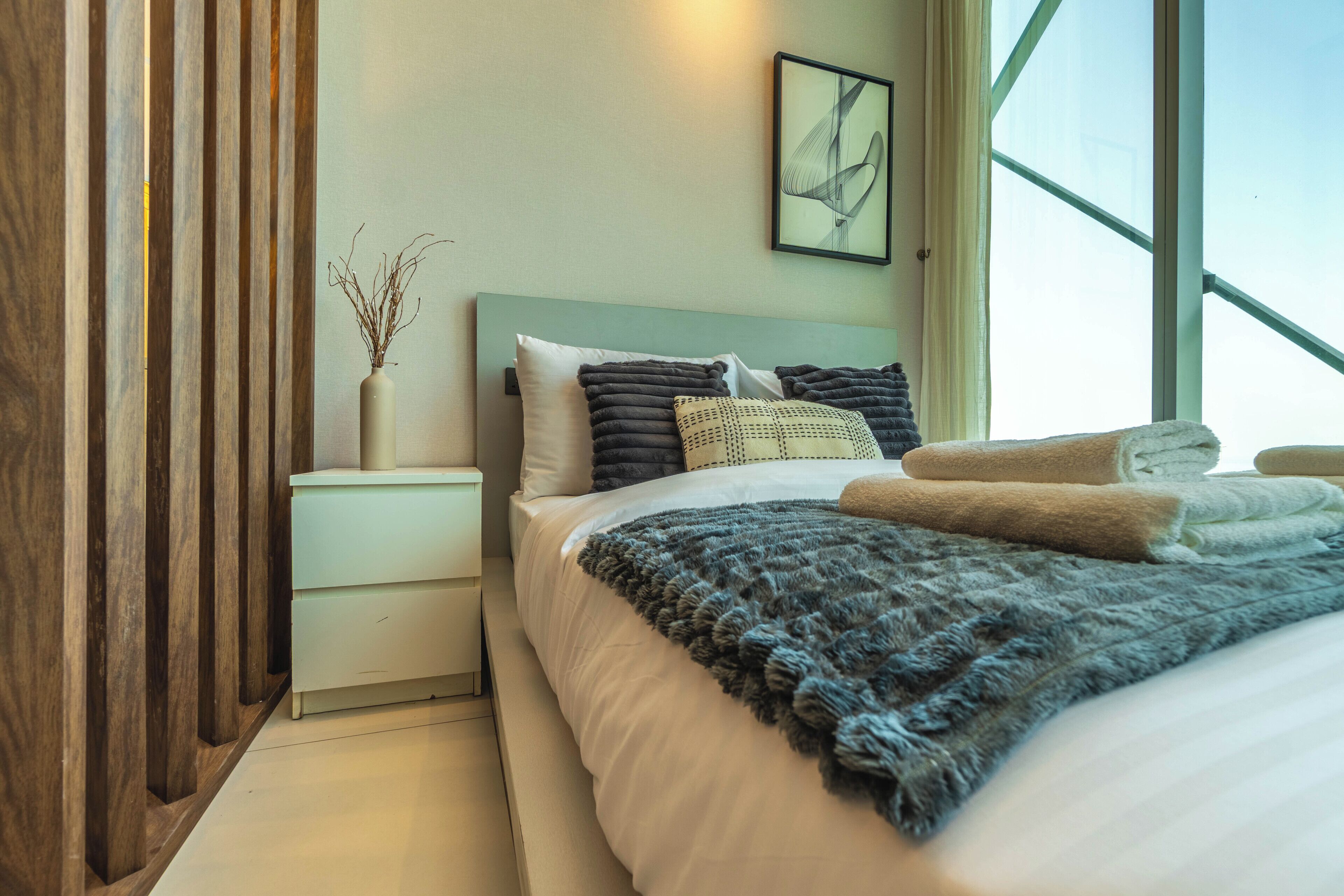 A modern bedroom with a large bed, a nightstand, a vase with branches, and a window with a view.