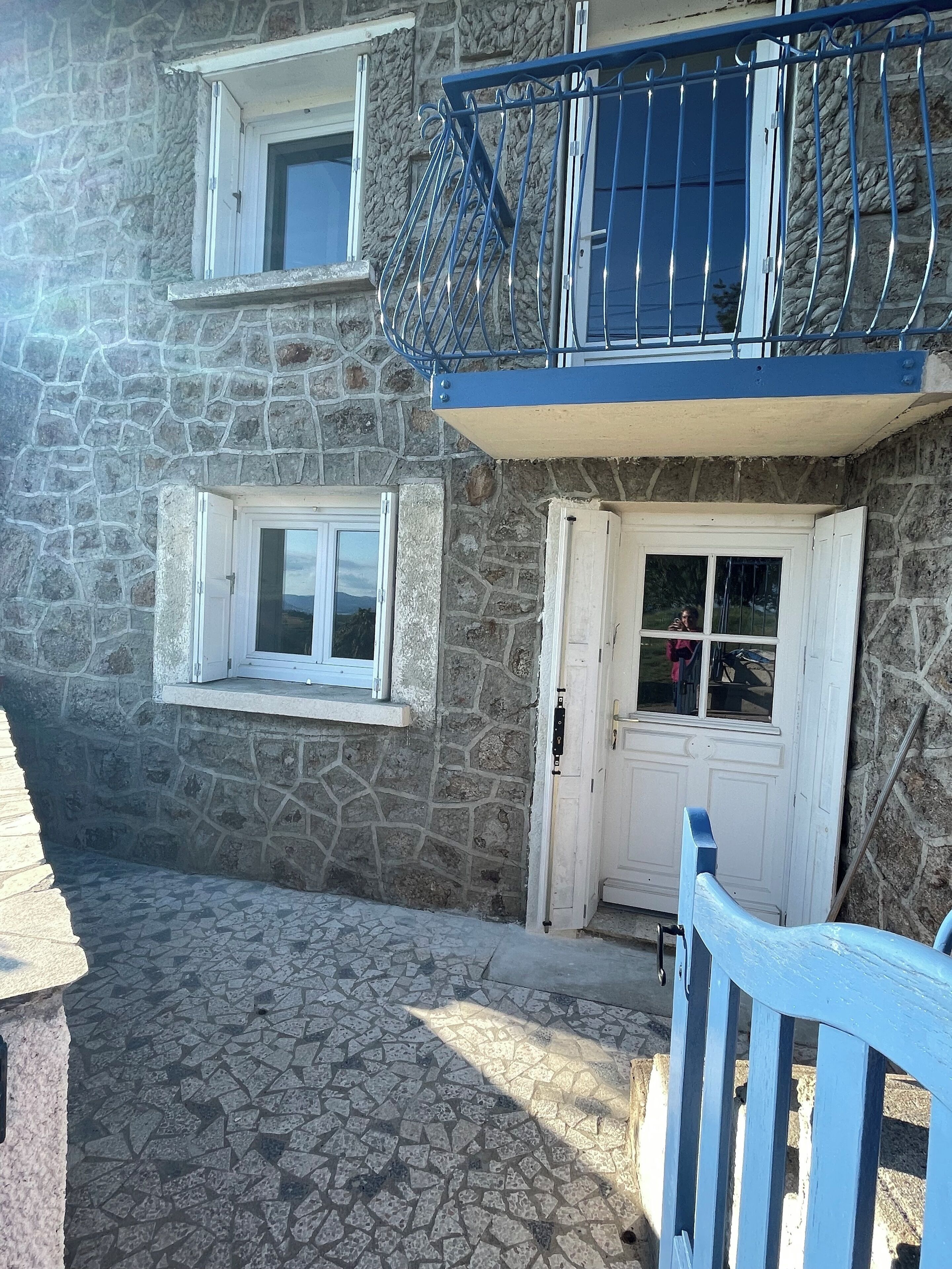 Ein Steinhaus mit einer weißen Tür und Fenstern, einem Balkon mit blauen Geländern und einem mit Fliesen gepflasterten Innenhof.