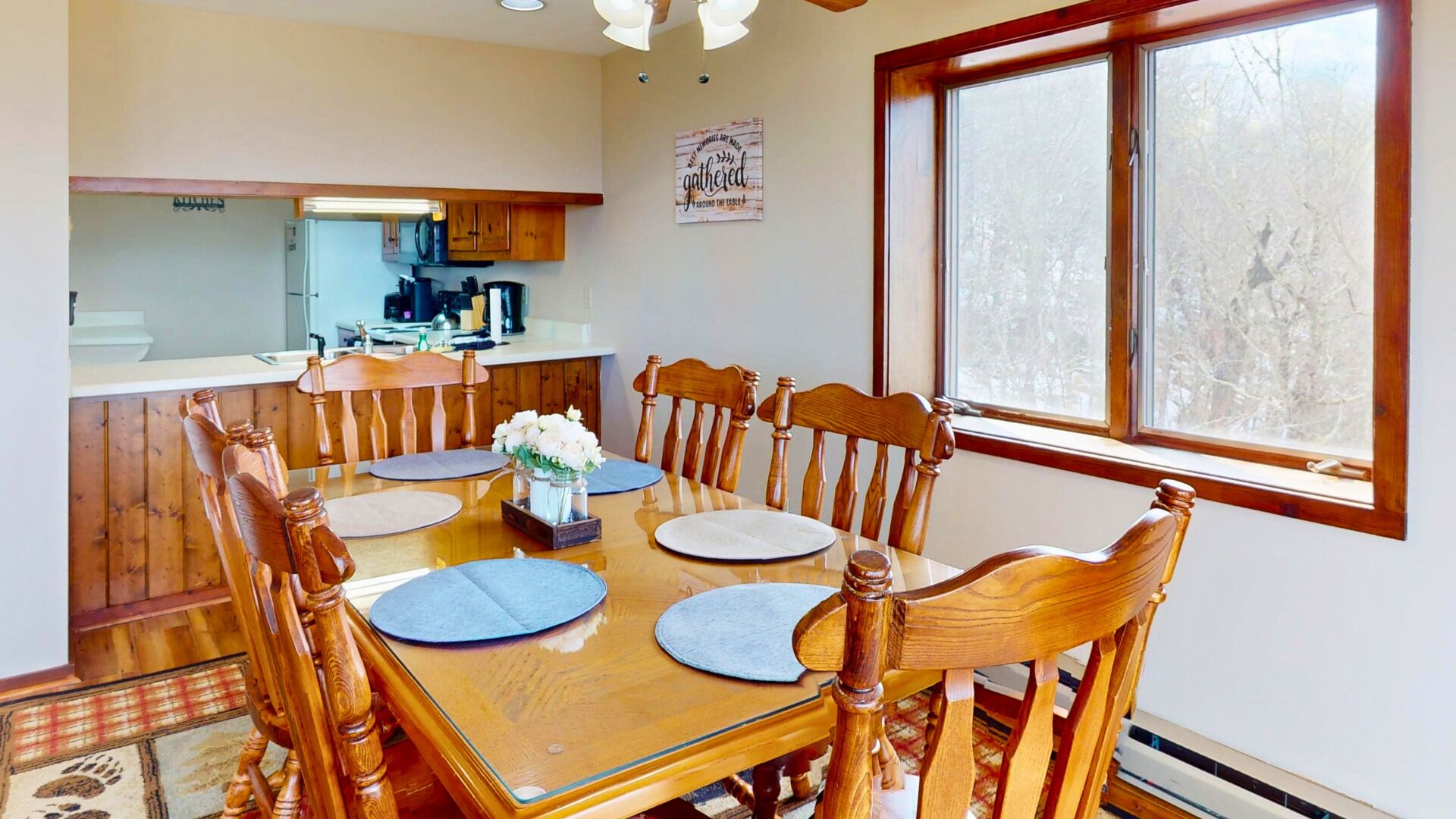 A dining area with a wooden table and chairs, a kitchen in the background, and a window with a view of trees.