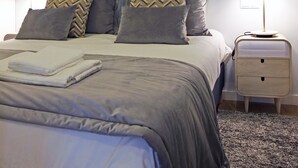 Cama bem arrumada com roupa de cama cinza e almofadas estampadas, um espelho fixado na parede, um abajur e uma pequena mesa de cabeceira.