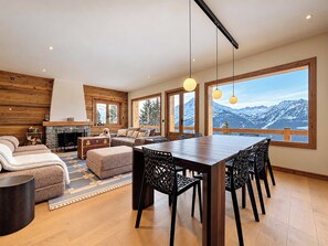 Amplia sala de estar con chimenea, un sofá, una mesa de comedor con sillas y ventanas grandes que ofrecen vistas a la montaña.
