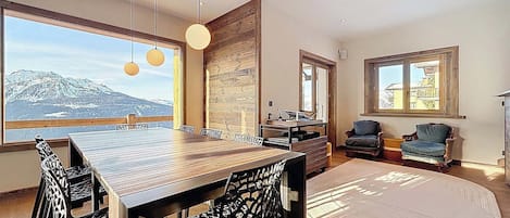 A modern dining area with a wooden table and chairs, large windows offering a mountain view, and a cozy seating area with two chairs.