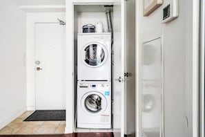 A laundry room with a front-loading washer and dryer, a small shelf with cleaning supplies, and a door leading to another room. - Executive Downtown Toronto Condo With A View (Toronto)