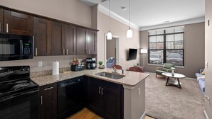 A modern kitchen with dark wood cabinets, a built-in microwave, and a granite countertop. There is a dining area with a round table and chairs, and a large window with blinds.