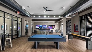 A pool table in a modern indoor space with a bar area, multiple screens, and a ceiling fan.