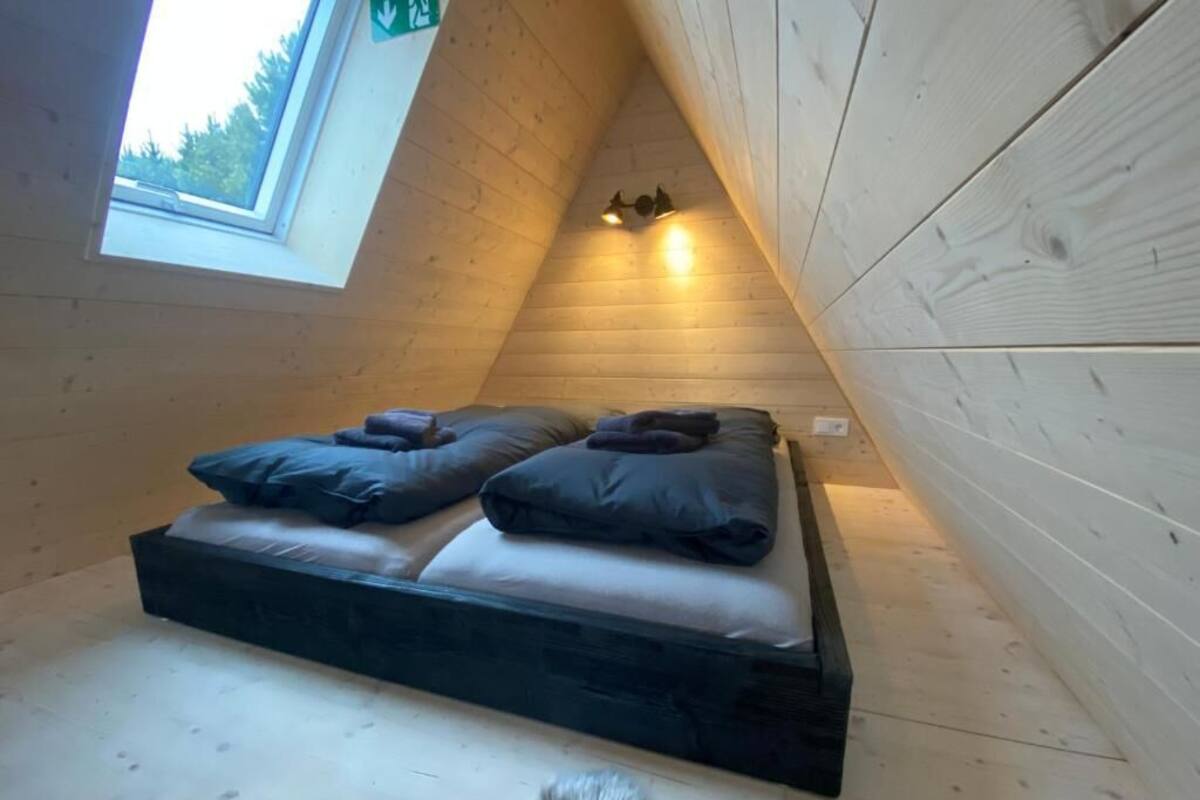 Une petite pièce triangulaire en bois, avec un lit, une lucarne et un luminaire fixé au mur.