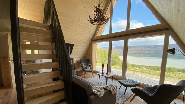 A modern interior with a wooden staircase, a sofa, two chairs, and a large window offering a view of a lake. - Hengifosslodge Tiny Houses (Egilsstadír)