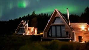 Two wooden cabins with pitched roofs, set against a backdrop of the Northern Lights. - Hengifosslodge Tiny Houses (Egilsstadír)