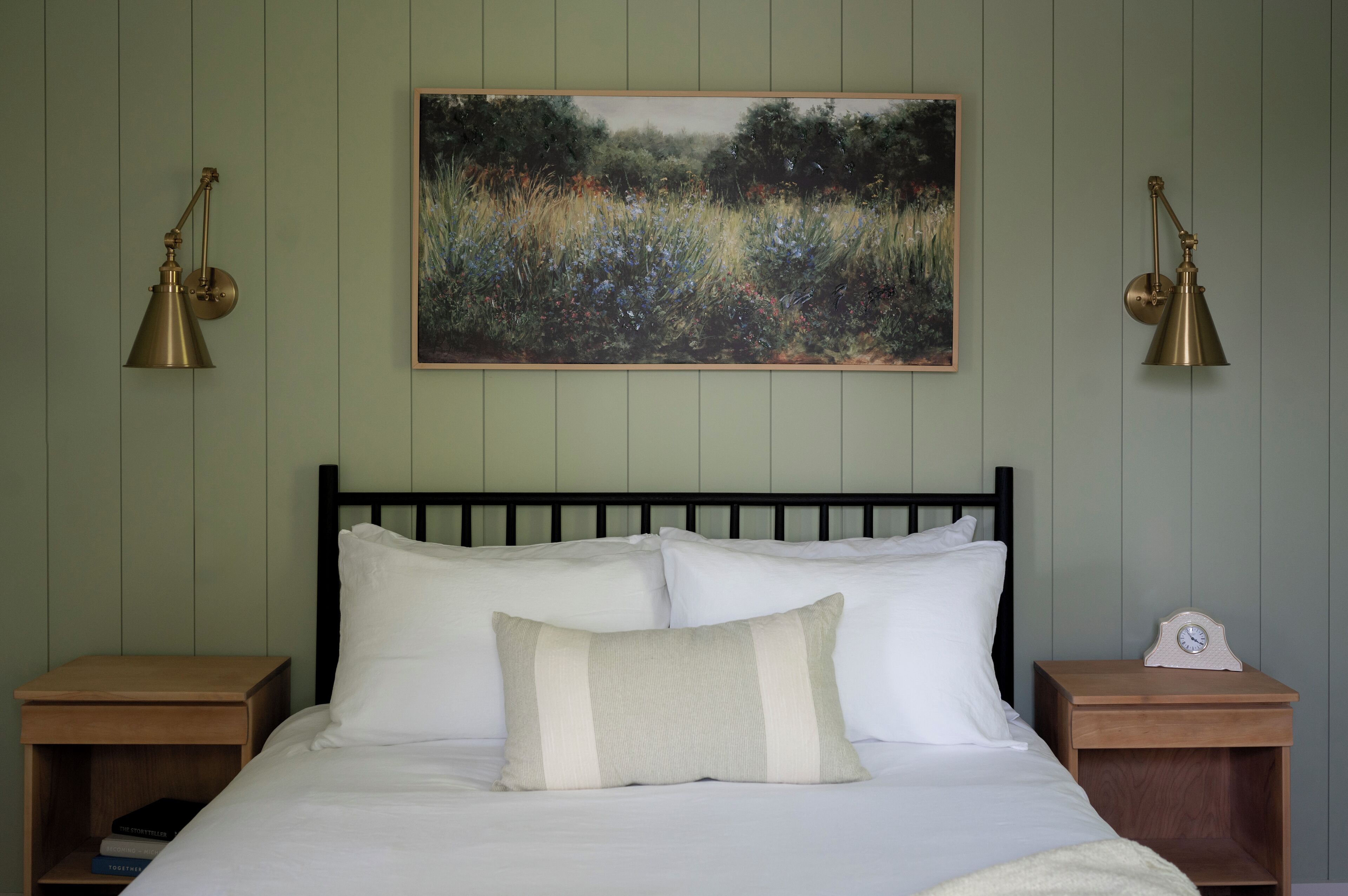 Una cama bien hecha con sábanas blancas y una única almohada decorativa, con dos mesitas de noche de madera, una de ellas con un pequeño reloj.