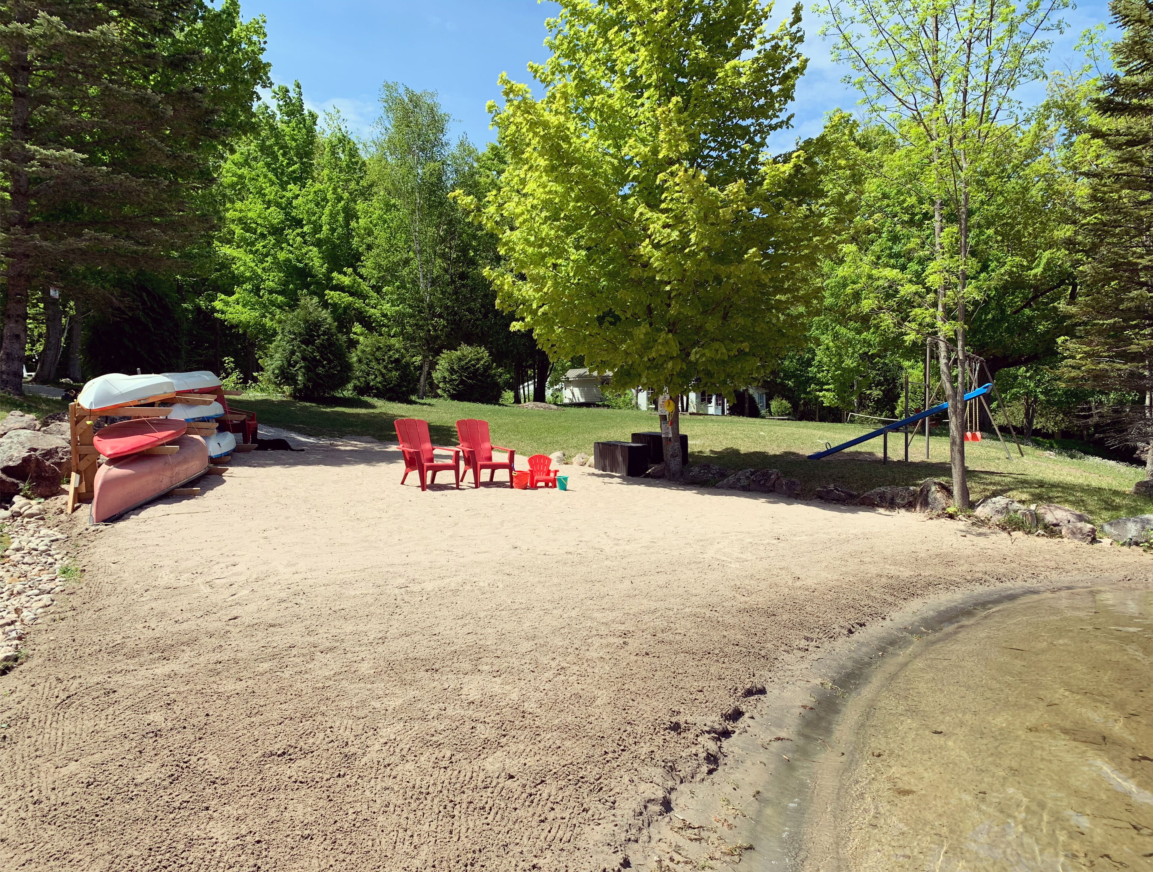 Une plage de sable avec des chaises Adirondack rouges, un canoë et une balançoire.