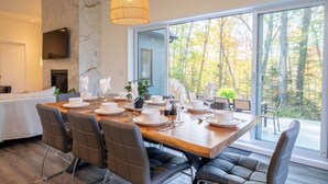 A dining area with a large table set for six, a fireplace, and a large window with a forest view.