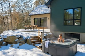 Une maison moderne avec un jacuzzi dans la neige, une terrasse avec des chaises et une grande fenêtre.
