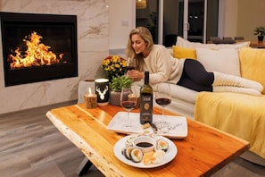 Un salon chaleureux avec une cheminée, un canapé, une table basse en bois sur laquelle sont disposés des sushis et une bouteille de vin, ainsi qu’une plante décorative.