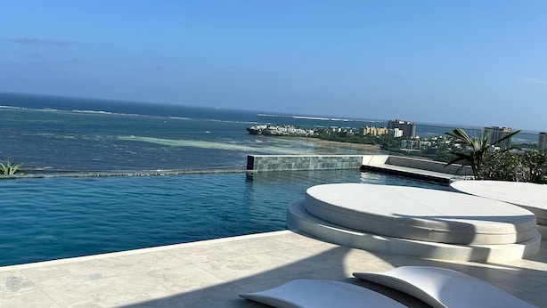 Area piscina moderna all'aperto con lettini, vista libera sull'oceano e skyline sullo sfondo.