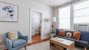 Un salon avec un fauteuil bleu, un canapé gris agrémenté de coussins orange et à motifs, un lampadaire blanc et une table basse sur laquelle repose un livre.