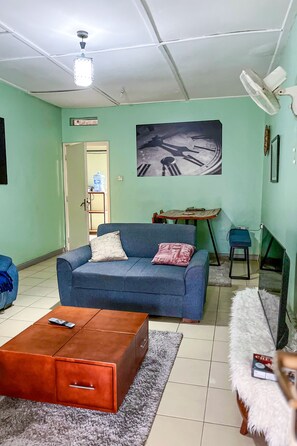 A living room with a blue sofa, a wooden coffee table, a white furry ottoman, and a ceiling fan. - Charming 2-bedroom apartment in enjoyable Kampala neighborhood free wifi (Kampala)