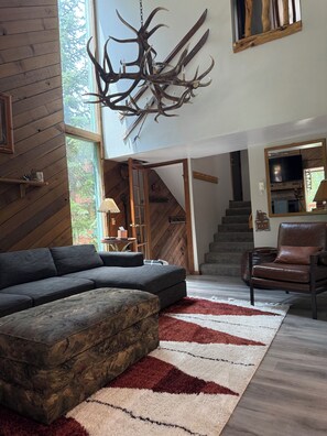 Un salon moderne avec un canapé d’angle, un pouf à motifs, un grand tapis, un mur en bois et un lustre original en forme de bois de cerf.