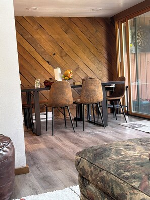 A dining area with a wooden wall, a table, chairs, and a view through a glass door.