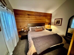 A bedroom with a wooden headboard, a bed with pillows, a nightstand with a lamp, and a window with curtains. - Mogul Manor-3B Giant Steps (Brian Head)