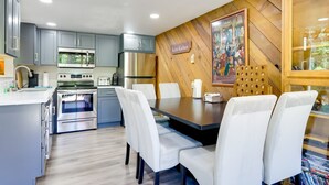 Una cocina moderna con armarios grises, electrodomésticos de acero inoxidable, una mesa de comedor con sillas blancas y una pared de acento de madera con un letrero que dice 'Scatter Kindness'.
