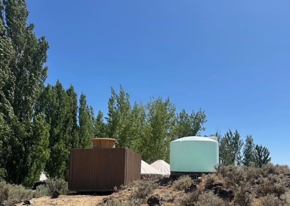 Dos estructuras cilíndricas grandes, posiblemente tanques de almacenamiento, en una zona árida y cubierta de arbustos, con árboles altos al fondo.